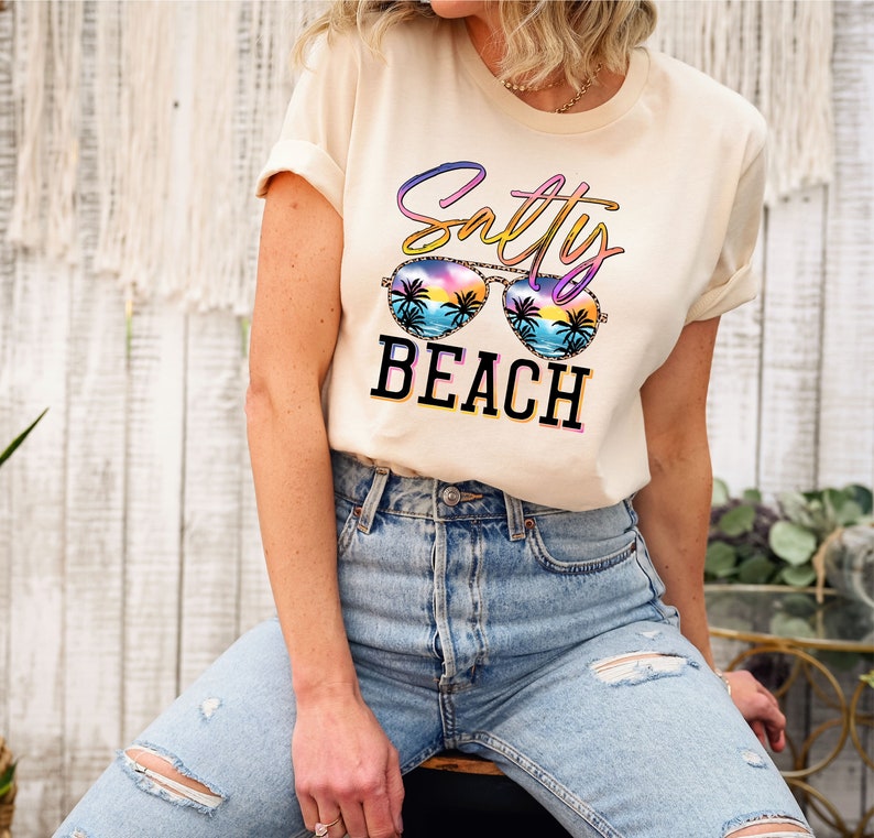 May include: A cream-colored t-shirt with the words "Salty Beach" in a colorful design. The sunglasses graphic features a beach scene with palm trees and a sunset. The shirt is paired with blue jeans.