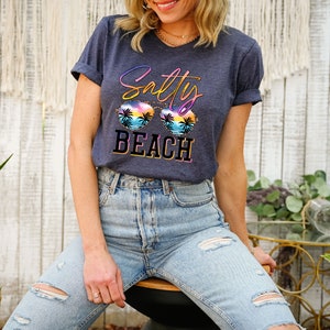 May include: A navy blue t-shirt with the words "Salty Beach" in a colorful design. The graphic includes sunglasses with a beach scene. The shirt is worn with light blue jeans.