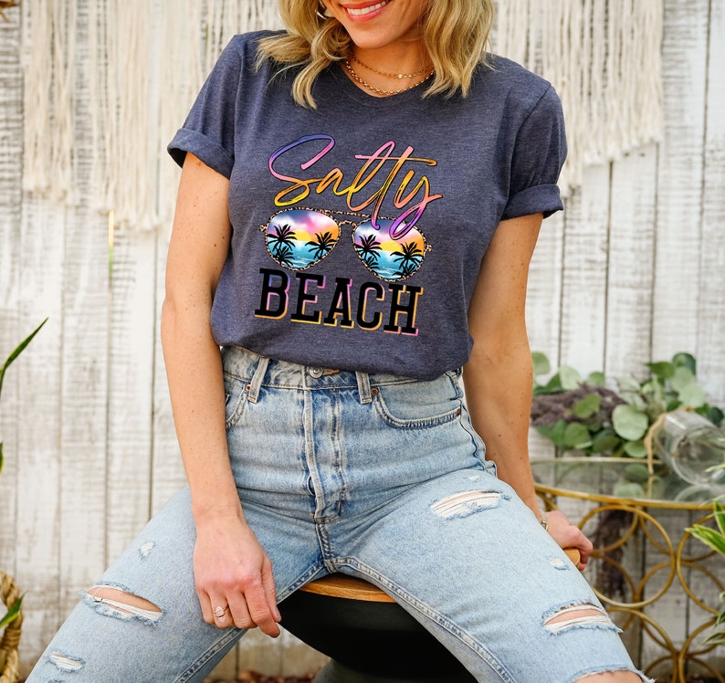 May include: A navy blue t-shirt with the words "Salty Beach" in a colorful design. The graphic includes sunglasses with a beach scene. The shirt is worn with light blue jeans.