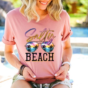 May include: A dusty rose t-shirt with the words "Salty Beach" in a colorful, stylized font. The design includes sunglasses with a beach scene inside. The shirt has rolled-up sleeves.