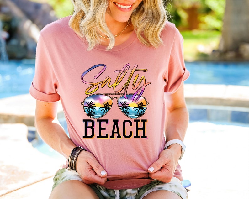 May include: A dusty rose t-shirt with the words "Salty Beach" in a colorful, stylized font. The design includes sunglasses with a beach scene inside. The shirt has rolled-up sleeves.