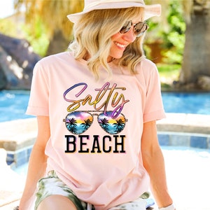May include: A light pink t-shirt with the words "Salty BEACH" in a colorful design. The word "Salty" is in a gradient of colors, and the word "BEACH" is in black block letters. The design includes sunglasses with a beach scene.