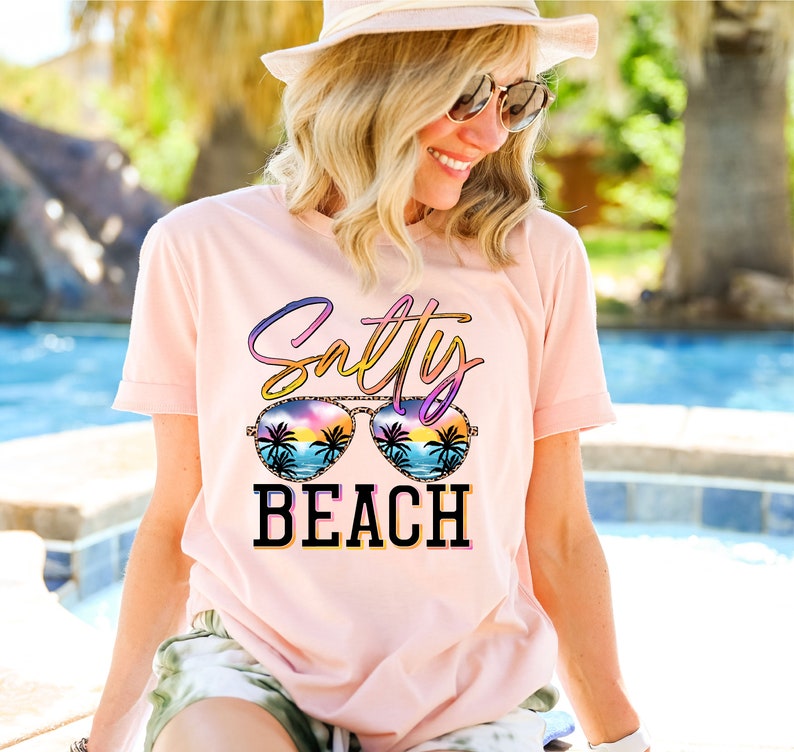 May include: A light pink t-shirt with the words "Salty BEACH" in a colorful design. The word "Salty" is in a gradient of colors, and the word "BEACH" is in black block letters. The design includes sunglasses with a beach scene.