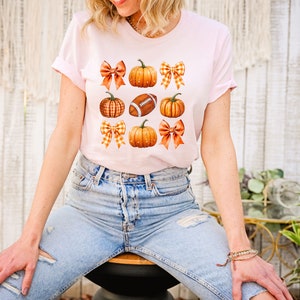 May include: A pink t-shirt with a graphic design of orange pumpkins, brown footballs, and orange and white gingham bows.