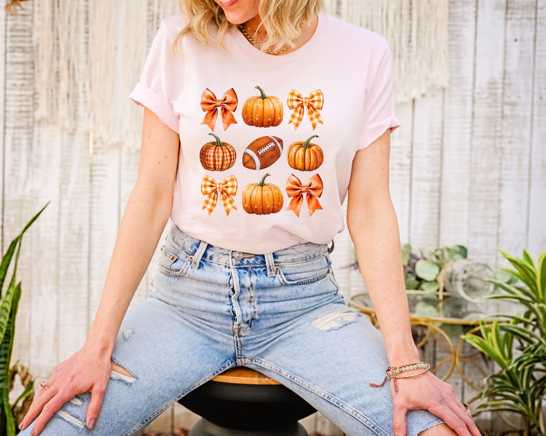 May include: A pink t-shirt with a graphic design of orange pumpkins, brown footballs, and orange and white gingham bows.