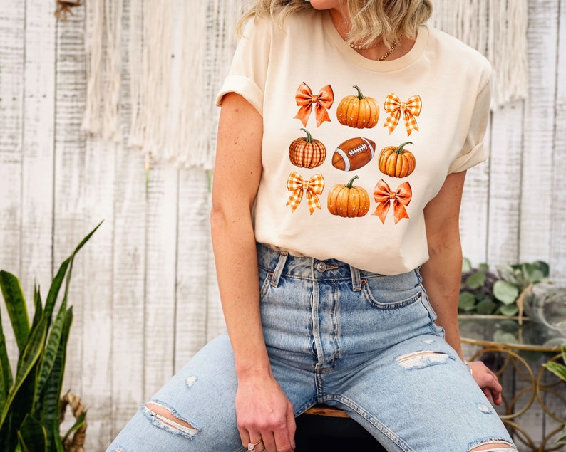 May include: A cream colored t-shirt with a graphic design of orange pumpkins, brown footballs, and orange and white checkered bows.