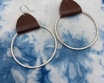 May include: A pair of silver hoop earrings with a hammered texture. Each earring features a brown leather accent at the top. The earrings are displayed against a blue and white tie-dye background.