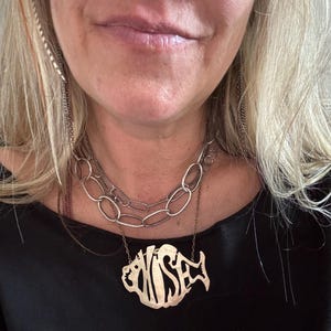 May include: A layered necklace set featuring a silver-toned chain with large oval links and a second, longer chain holding a gold-toned pendant shaped like a stylized fish with the letters "WISE" cut out from its body in a bold, decorative font.