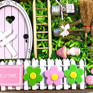 May include: A miniature fairy garden kit with a pink door, white picket fence, and colorful felt flowers. Includes a wooden ladder, tiny broom, and a welcome sign. The kit also features a small bottle labeled "Fairy Treasures".