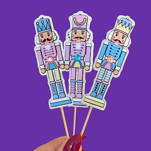 May include: Three colorful nutcracker cupcake toppers on wooden sticks. Each nutcracker has a different color scheme: pink, purple, and blue. The toppers have glitter accents and are held against a purple background.