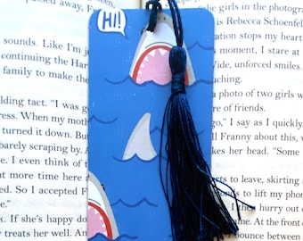May include: A blue bookmark with a white shark design and the words "Hi!" and "Hey!" in speech bubbles. The bookmark has a blue tassel.