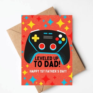 May include: A red card with a black and blue video game controller on it. The card has the text "LEVELED UP TO DAD!" and "HAPPY 1ST FATHER'S DAY!" on it. The background is a red and orange pattern with yellow and blue stars.
