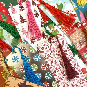 May include: A collection of Christmas-themed bookmarks with colorful tassel accents. The bookmarks feature various festive patterns, including Christmas trees, candy canes, holly, and gingerbread houses. Tassels come in red, pink, green, gold, and blue.