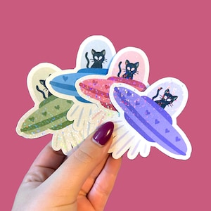 May include: A collection of colorful stickers featuring a black cat in a UFO. The stickers have a white border and are in shades of green, blue, pink, and purple. Each UFO has heart details and glitter accents. The background is a solid pink.