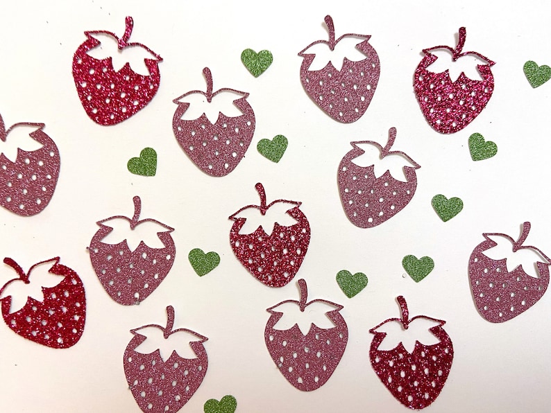 May include: A scattering of pink and red glitter-covered paper cutouts of strawberries and green hearts on a white background.