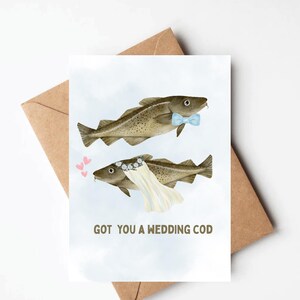 May include: A greeting card featuring two illustrated codfish, one wearing a blue bow tie and the other a bridal veil. The text "GOT YOU A WEDDING COD" is printed below the fish, with three pink hearts on the left.