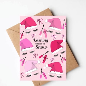 May include: A pink greeting card with a festive design. The card features illustrations of Santa hats, eyelashes, mascara wands, and bows. The text "Lashing Through the Snow" is displayed in the center. The card is placed on a brown envelope.