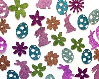 Puede incluir: Recortes brillantes con temática de Pascua en varias formas y colores. La imagen presenta flores moradas, verdes y marrones, huevos de Pascua turquesa y morados, y siluetas de conejos morados sobre una superficie blanca.