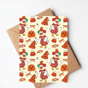 May include: A greeting card with a repeating pattern of a clown duck, pumpkins, candy corn, and party hats on a cream background. The clown duck is wearing a red and white striped shirt and a clown hat. The card is placed on a brown envelope.