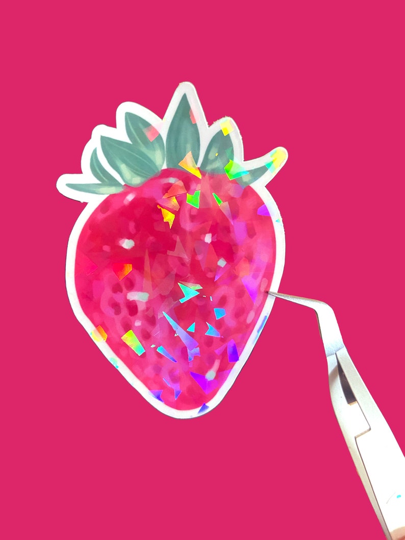 Puede incluir: Una pegatina hologr&aacute;fica de una fresa roja con hojas verdes. La pegatina est&aacute; sobre un fondo rosa y se sostiene con unas pinzas.