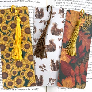 May include: Three bookmarks with different designs. The first bookmark features a sunflower pattern with a yellow tassel. The second bookmark has a brown tassel and a pattern of squirrels and acorns. The third bookmark has a gold tassel and a pattern of autumn leaves in shades of red, orange, and yellow.