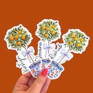 May include: Watercolor stickers featuring miniature orange trees in blue and white patterned pots. Each tree has a green leafy canopy with orange fruit and a light blue bow. The names "Kirstyn", "Jovana", and "Rishab" are written on the stickers.