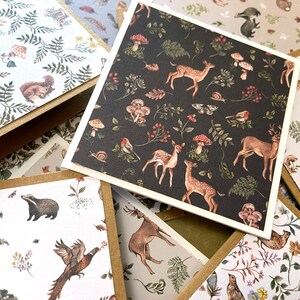 May include: A collection of square cards with woodland-themed illustrations. One card features a dark background with deer, mushrooms, and birds. Other cards have similar designs with squirrels, ferns, and other forest creatures. The cards have a white border and are set against a brown background.