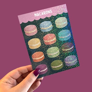 May include: A card featuring colorful macarons. The card has a dark green glittery background and a pink scalloped border at the top with the word "MACARONS" printed in white. The macarons are in various colors, including pink, blue, and green.