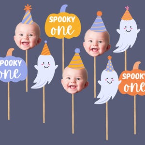 May include: A collection of Halloween-themed photo booth props. The props include baby faces wearing party hats, ghost cutouts, and pumpkin cutouts. The pumpkins and ghosts have the words "Spooky One" written on them. The props are on wooden sticks.