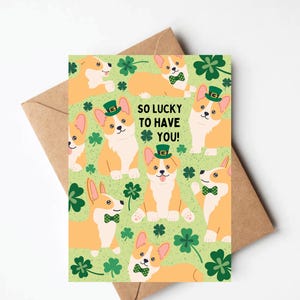 May include: A green and white card with a pattern of orange corgis wearing green hats and bowties. The text "SO LUCKY TO HAVE YOU!" is in black letters.