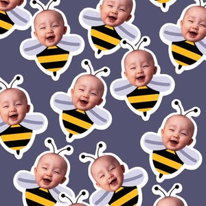 May include: A repeating pattern of smiling baby faces superimposed on cartoon bee bodies. The bees have black and yellow striped bodies, white wings, and antennae. The background is a solid, muted purple.