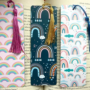 May include: Four colourful bookmarks with rainbow patterns and tassels. The bookmarks are on a book with text. The bookmarks are made of paper and have a white background with colourful rainbows and clouds. The tassels are pink, gold, blue, and white.