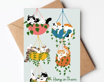 May include: A greeting card with a light blue background featuring five cats hanging in colorful plant pots. The cats are in various poses, some sleeping, some looking at the viewer. The text "Hang in There" is printed in black at the bottom of the card.