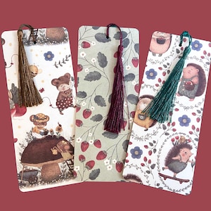 May include: Three illustrated bookmarks with colorful tassels. The bookmarks feature whimsical forest scenes with animals, including bears and hedgehogs. One bookmark has a brown tassel, one has a burgundy tassel, and one has a green tassel.