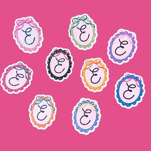 May include: A collection of nine oval stickers with a decorative border and a bow at the top. Each sticker features the letter "E" in a cursive font. The stickers have different color schemes, including pink, green, black, orange, purple, and blue.