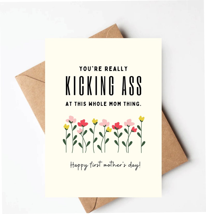 May include: A greeting card with the text "YOU'RE REALLY KICKING ASS AT THIS WHOLE MOM THING." and "Happy first mother's day!" in black font. The card features a floral design with pink, red, and yellow flowers. The card is on a brown envelope.