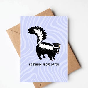 May include: A graduation greeting card featuring a cartoon skunk wearing a mortarboard. The skunk is black and white, set against a light blue background with wavy lines. The text "SO STINKIN' PROUD OF YOU" is below the skunk. The card is paired with a brown envelope.