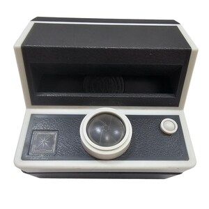 Op de afbeelding: Een vintage zwart-wit speelgoedcamera met een textuuroppervlak. De camera heeft een lens, een knop en een decoratief vierkant. Het bovenste gedeelte is zwart en de basis is wit.