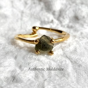 May include: A gold ring with a raw, green moldavite stone set in the center. The ring is made of a thin, hammered band. "Authentic Moldavite" is written below the ring.