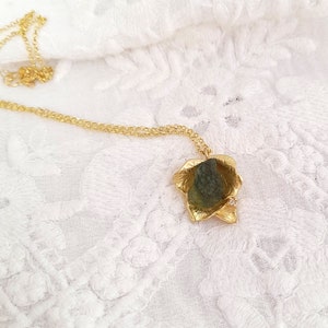 以下が含まれることがあります： 金色のネックレス。葉の形をしたペンダントが付いています。ペンダントには緑色の石と小さなダイヤモンドが入っています。繊細なチェーンは、白くテクスチャ加工された表面に置かれています。