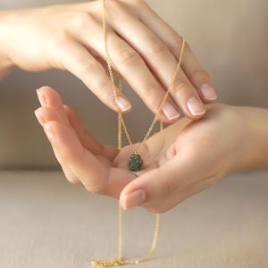 May include: A delicate gold chain necklace featuring a small, textured, dark olive-green raw stone pendant. The thin metallic chain links are uniform in size, supporting the rough, uneven surface of the central gemstone which hangs centrally.