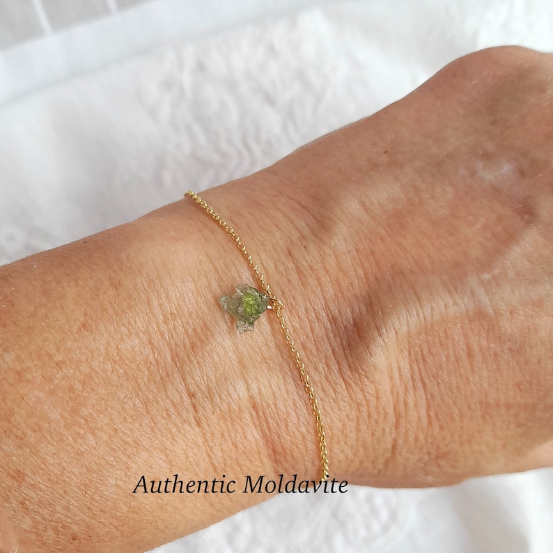 K&ouml;nnte beinhalten: Ein goldenes Kettenarmband mit einem gr&uuml;nen Moldavit-Anh&auml;nger. Der Text "Authentic Moldavite" ist unterhalb des Armbands sichtbar.