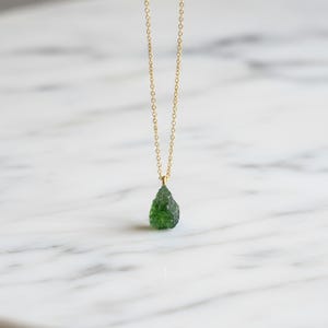 May include: A delicate gold chain necklace featuring a single, raw, teardrop-shaped green gemstone pendant. The pendant has a textured, uneven surface and is suspended from a thin, polished gold-tone metal link chain that hangs vertically.