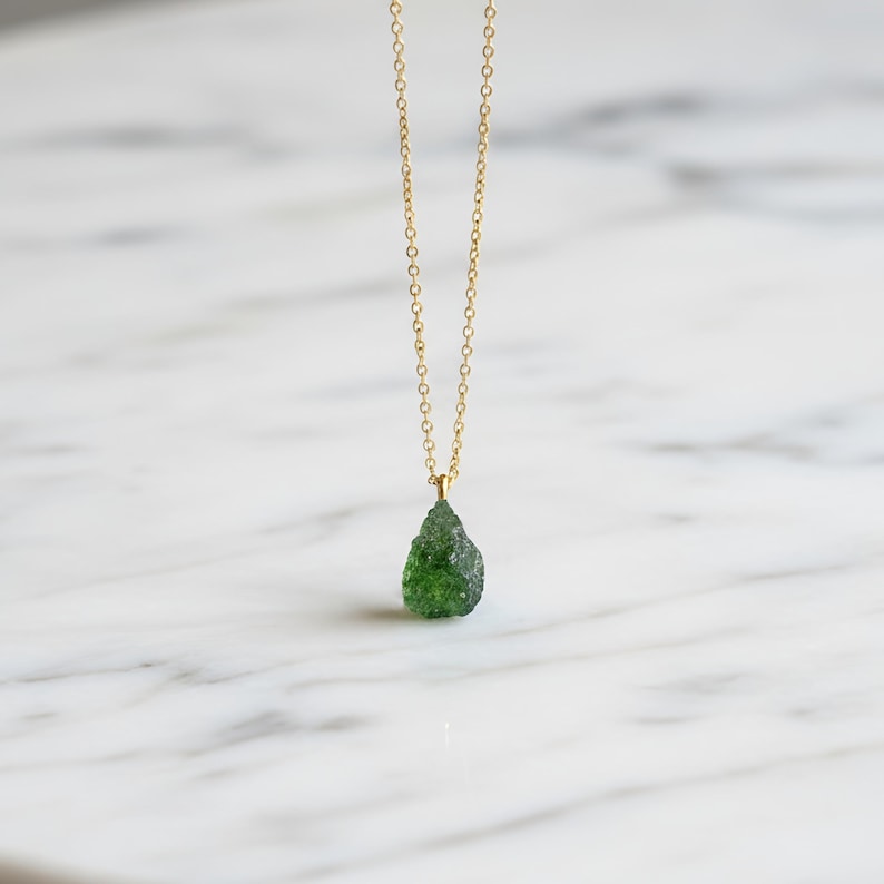 May include: A delicate gold chain necklace featuring a single, raw, teardrop-shaped green gemstone pendant. The pendant has a textured, uneven surface and is suspended from a thin, polished gold-tone metal link chain that hangs vertically.