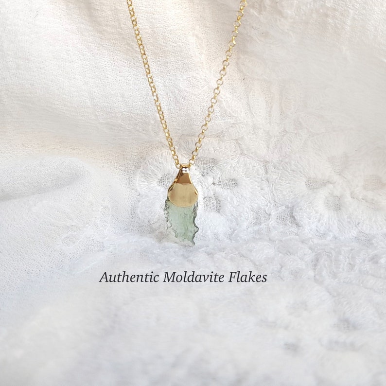 May include: A gold chain necklace with a pendant featuring a green moldavite flake set in a gold bezel. The pendant is on a white fabric background. Authentic Moldavite Flakes.