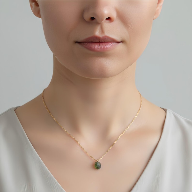 May include: A delicate gold chain necklace featuring a small, raw, textured green gemstone pendant. The necklace rests against the neck, showcasing a thin, polished gold link chain that holds the irregular, dark olive-green stone securely.