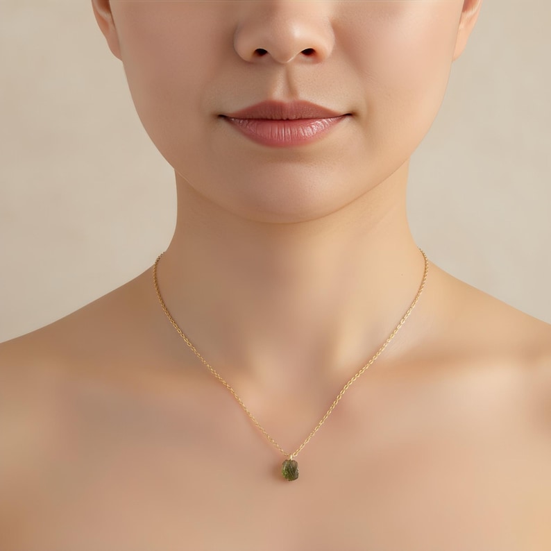May include: A delicate gold chain necklace featuring a small, raw, olive-green gemstone pendant. The necklace rests against the neck, showcasing a thin, polished metal chain that holds the textured, irregular stone in a simple, centred setting.