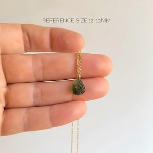 May include: A necklace featuring a small, rough, olive-green stone pendant suspended from a thin gold-coloured chain. The text REFERENCE SIZE 12-13MM is visible above the pendant, indicating the approximate size of the stone in millimetres.