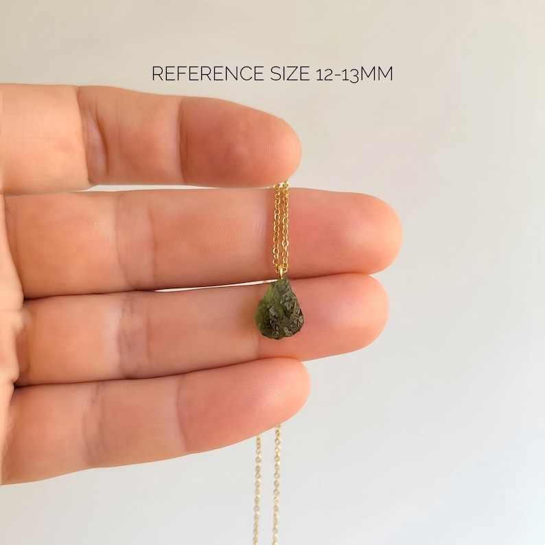 May include: A necklace featuring a small, rough, olive-green stone pendant suspended from a thin gold-coloured chain. The text REFERENCE SIZE 12-13MM is visible above the pendant, indicating the approximate size of the stone in millimetres.