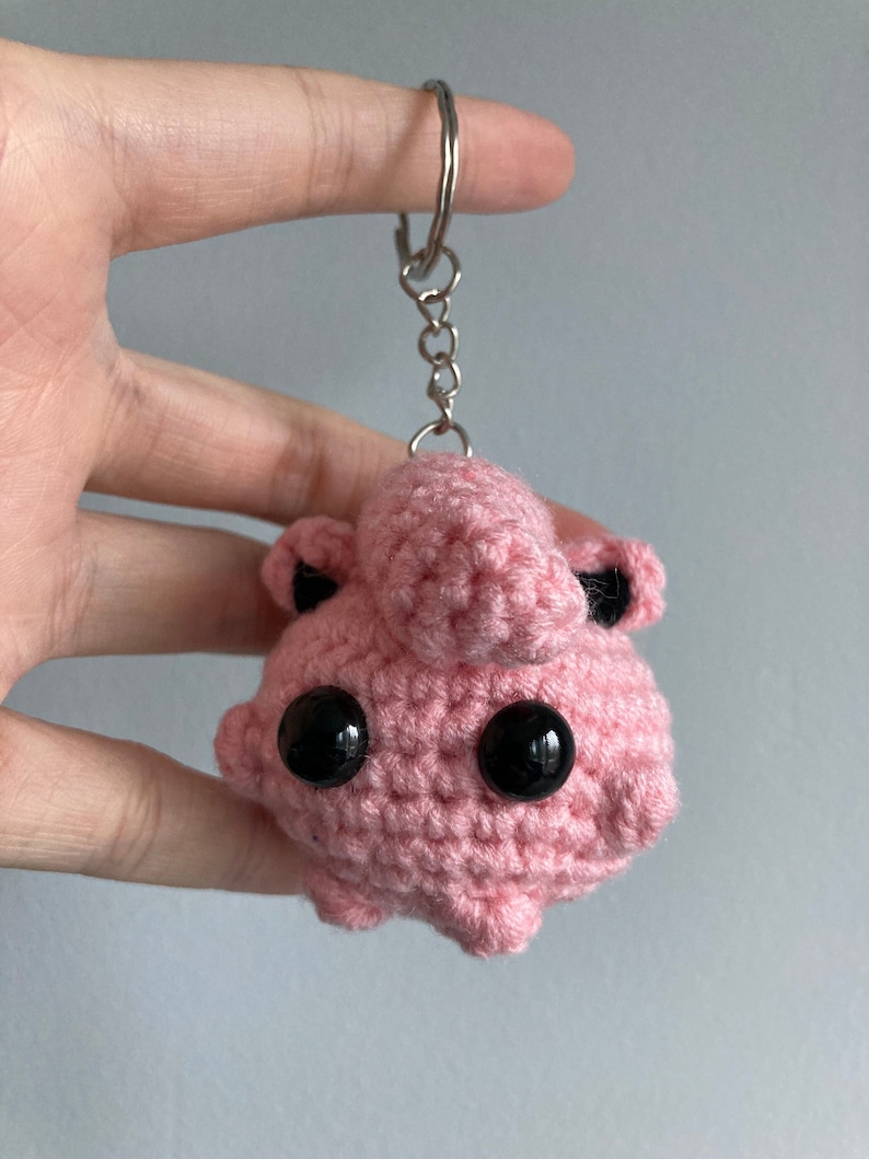 May include: Pink crocheted keychain with black eyes and a silver chain. The keychain is shaped like a cartoon character.